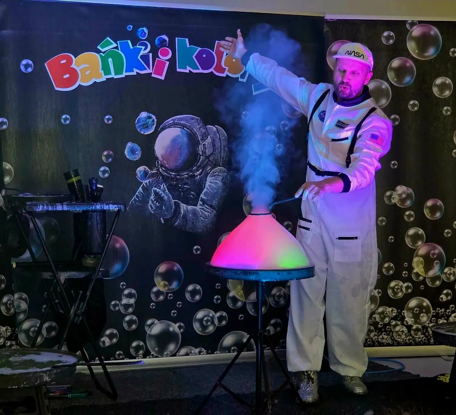 Bańka wypełniona kolorowym dymem na scenie, prowadzący w kombinezonie astronauty